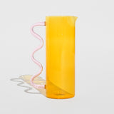 Photo of the Wave Pitcher - Yellow/Pink, a tall glass pitcher with a yellow cylindrical tank and a pink wavy glass handle.
